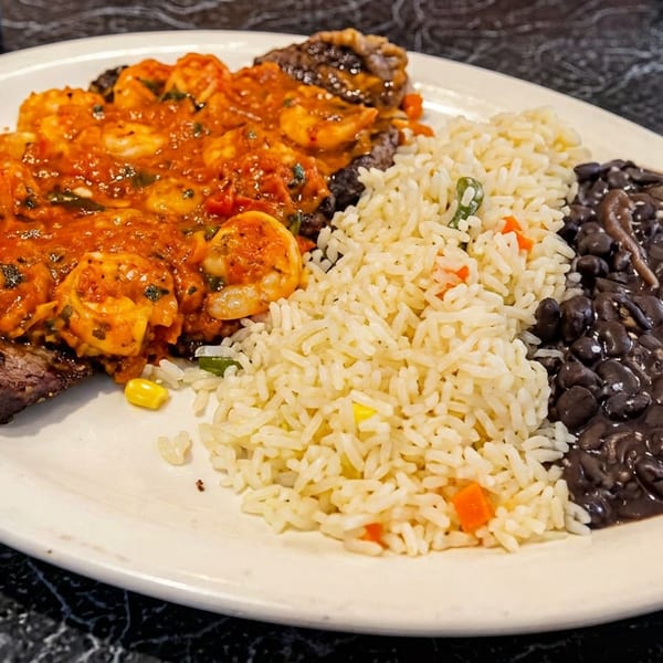 Steak topped with shrimp in red sauce, served with rice and black beans on a white plate.